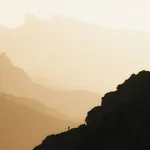 Eine weite Berglandschaft, die in ein sanftes, goldenes Licht getaucht ist, mit einer einsamen Gestalt, die auf einem Felsvorsprung steht und in die Ferne blickt.