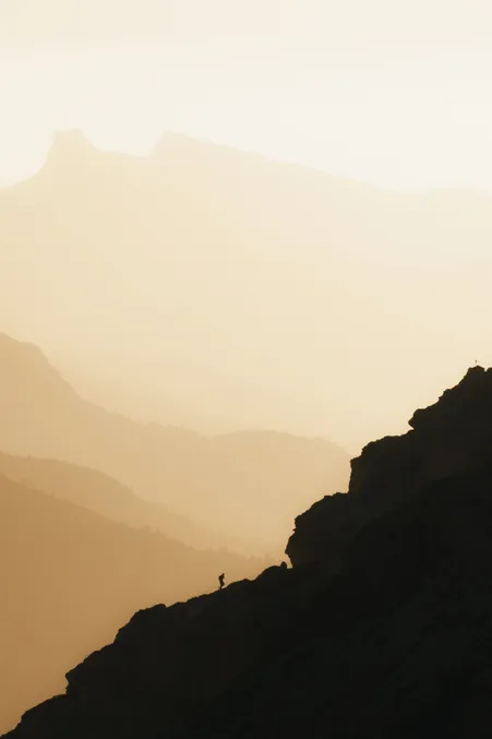 Eine weite Berglandschaft, die in ein sanftes, goldenes Licht getaucht ist, mit einer einsamen Gestalt, die auf einem Felsvorsprung steht und in die Ferne blickt.